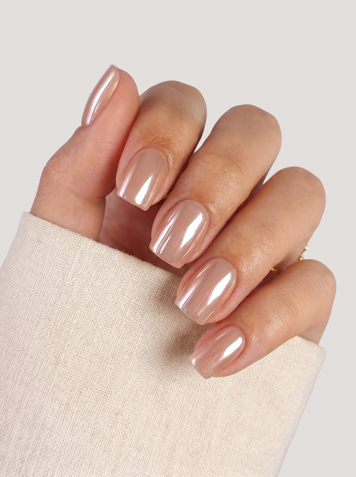 A close-up of a hand with a glossy, metallic nude-pink manicure, wearing a cream-colored knit sleeve against a light gray background.